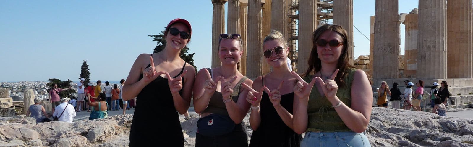 Four students make a "W" with their hands and stand in front of the ruined columns of the Parthenon